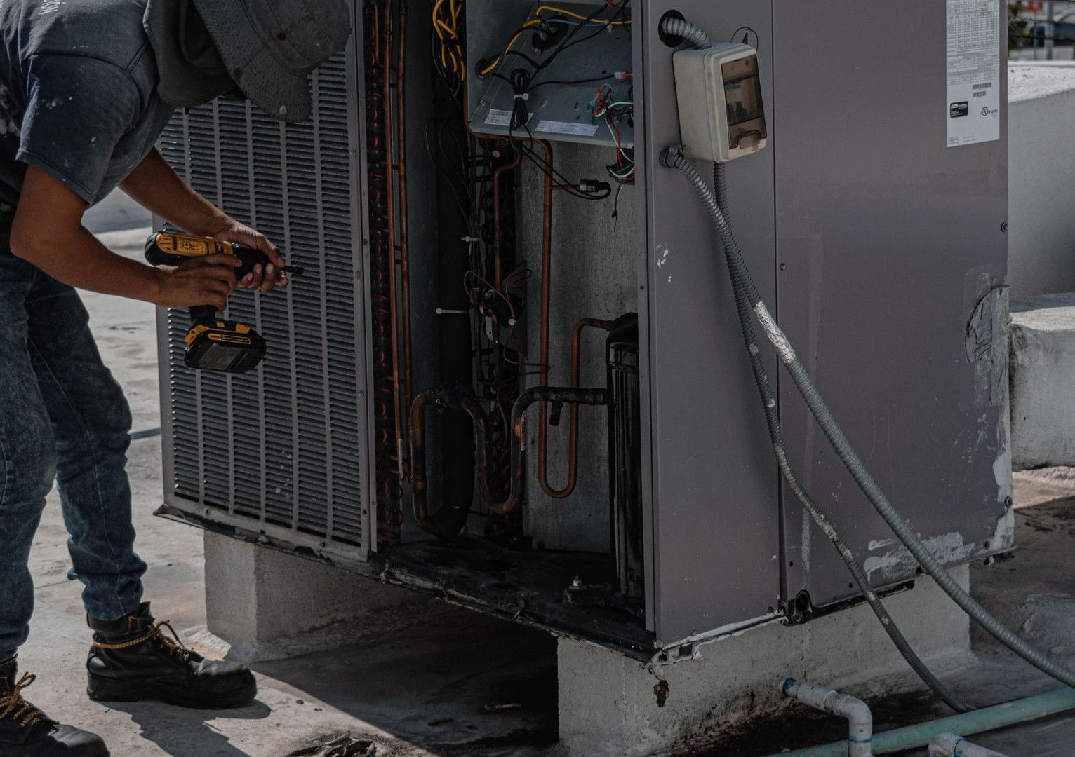 A worker in a bucket hat repairs an outdoor air conditioning unit on a rooftop.